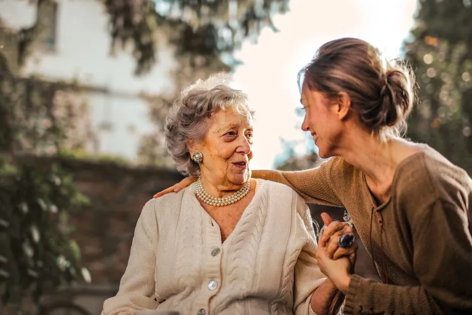 An older woman and an adult woman holding hands and smiling together outdoors, illustrating a caregiving relationship between generations.
