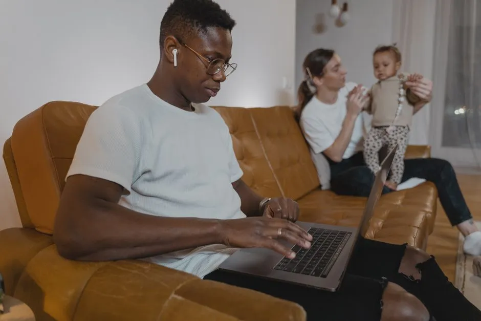 A parent working on a laptop on the sofa while another family member looks after a toddler nearby, illustrating the reality of working from home with young kids.